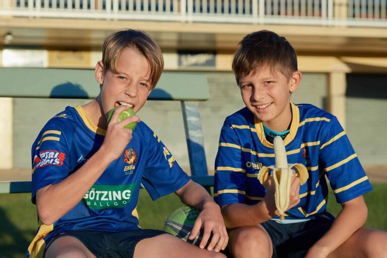 Two 14 year old boys sitting on concrete steps wearing a sport gernsey. One holds a peeled banana, the other is biting into a green apple.