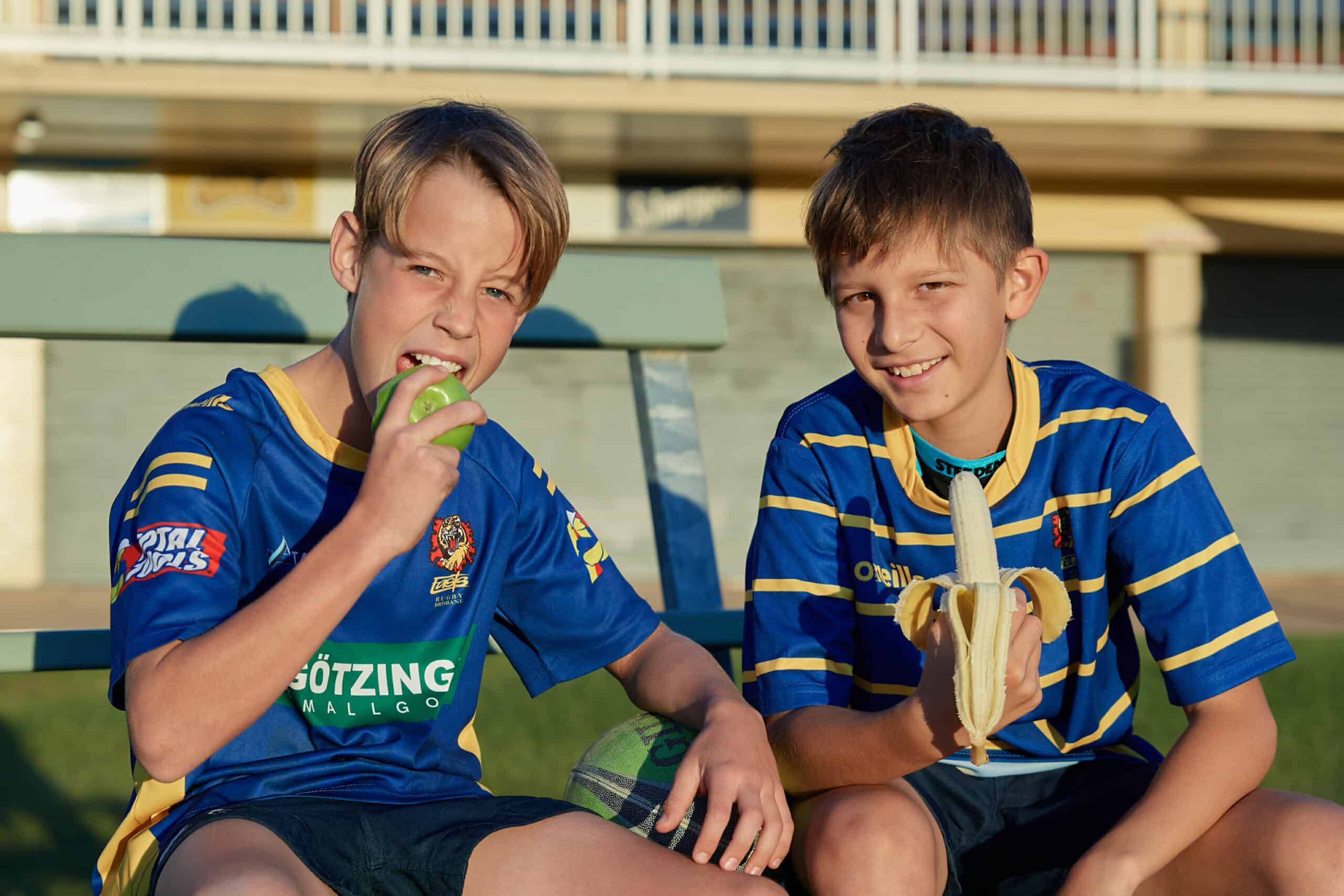 Two 14 year old boys sitting on concrete steps wearing a sport gernsey. One holds a peeled banana, the other is biting into a green apple.