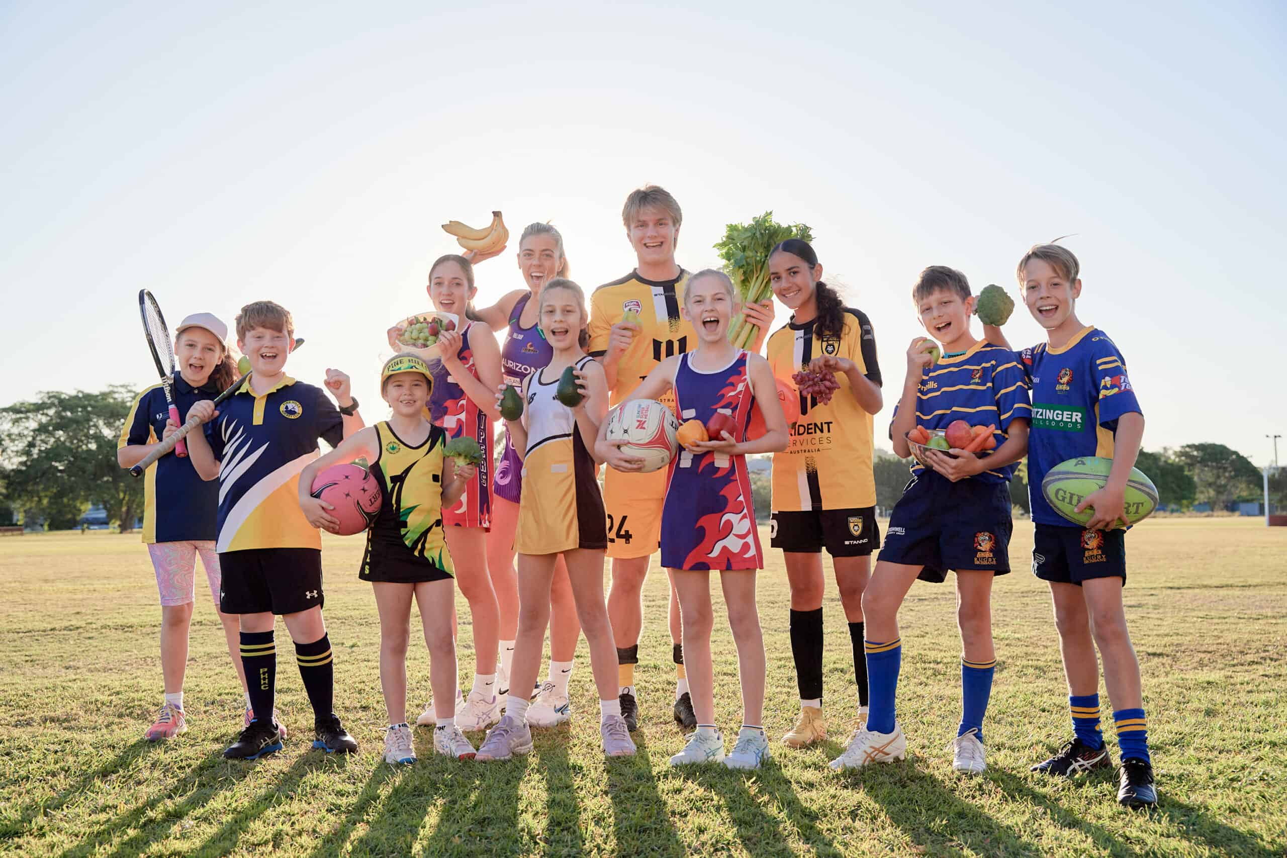 Group of 11 school students in sports wear standing on a grass oval holding fruit and vegetables and sports equipment