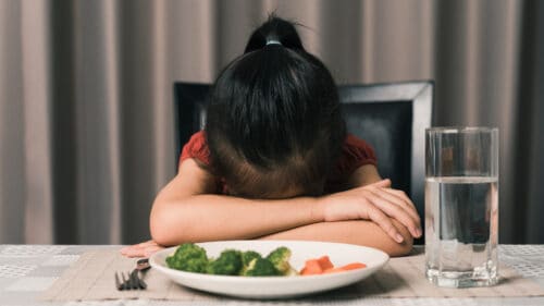 Child with head down on table, refusing to eat a plate of vegetables in front of them. 