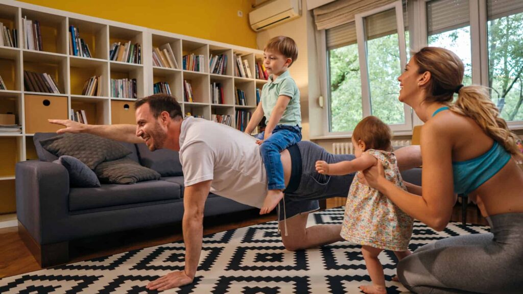 Father doing some yoga in the living room while son sits on his back and mother and daughter watch on laughing