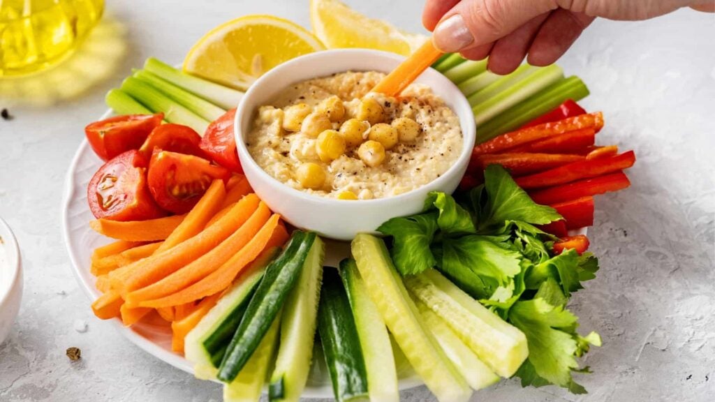 Vegetable platter featuring fresh carrot sticks, sliced cherry tomatoes, cucumber, sliced red capsicum, celery sticks and fresh lemon wedges, with a hummus dip in the centre