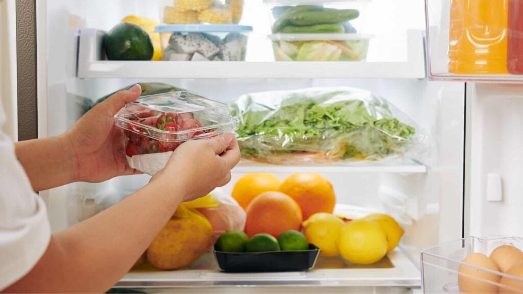 A person removing a packet of strawberries from a fridge that is full of vegetables and fruit.