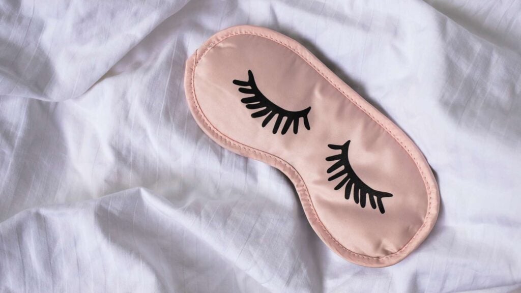 Pink silk sleeping mask with eyelashes printed on it rests on white bed sheet for article about a diet to help you sleep better