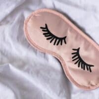 Pink silk sleeping mask with eyelashes printed on it rests on white bed sheet for article about a diet to help you sleep better