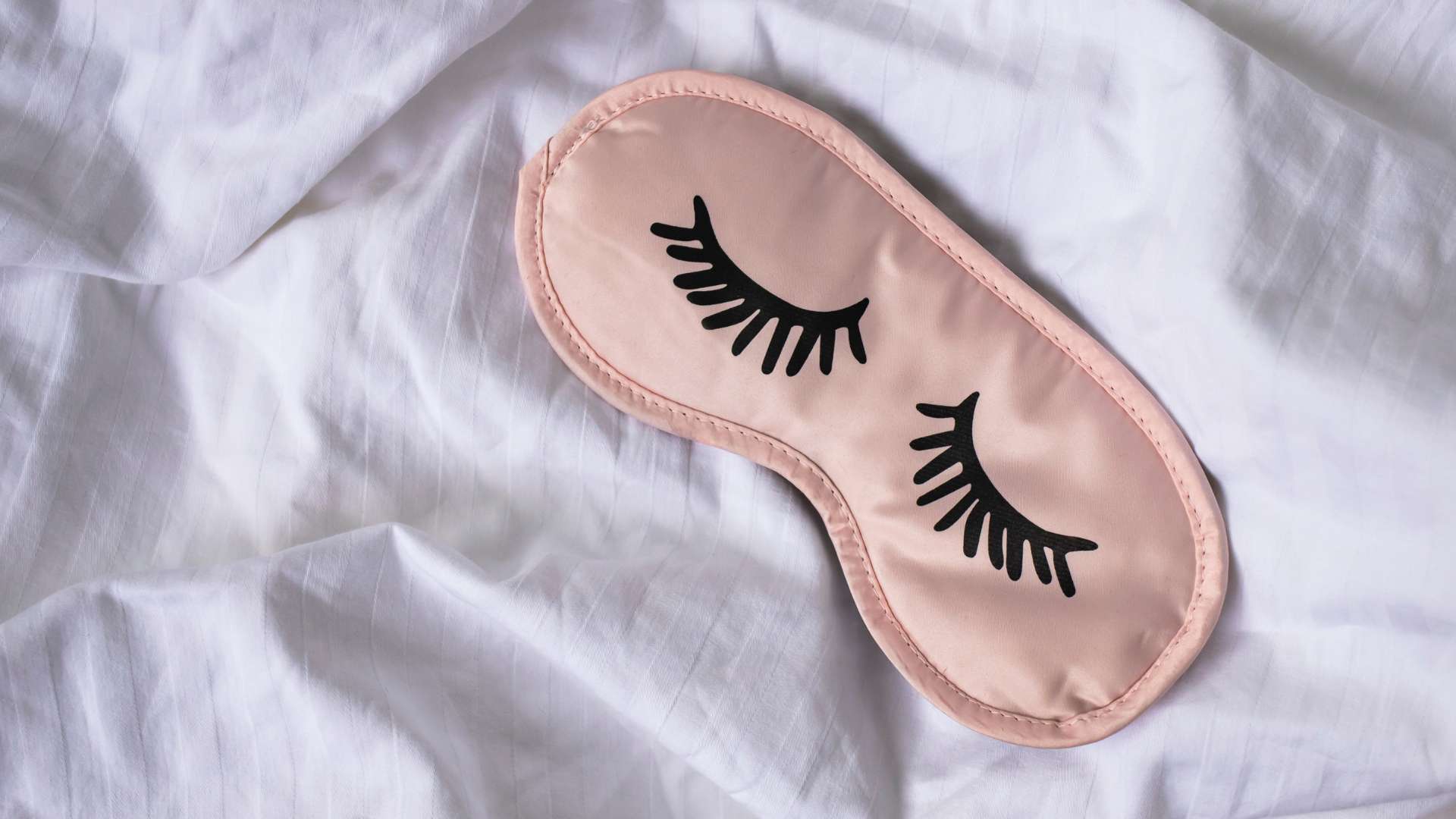 Pink silk sleeping mask with eyelashes printed on it rests on white bed sheet for article about a diet to help you sleep better