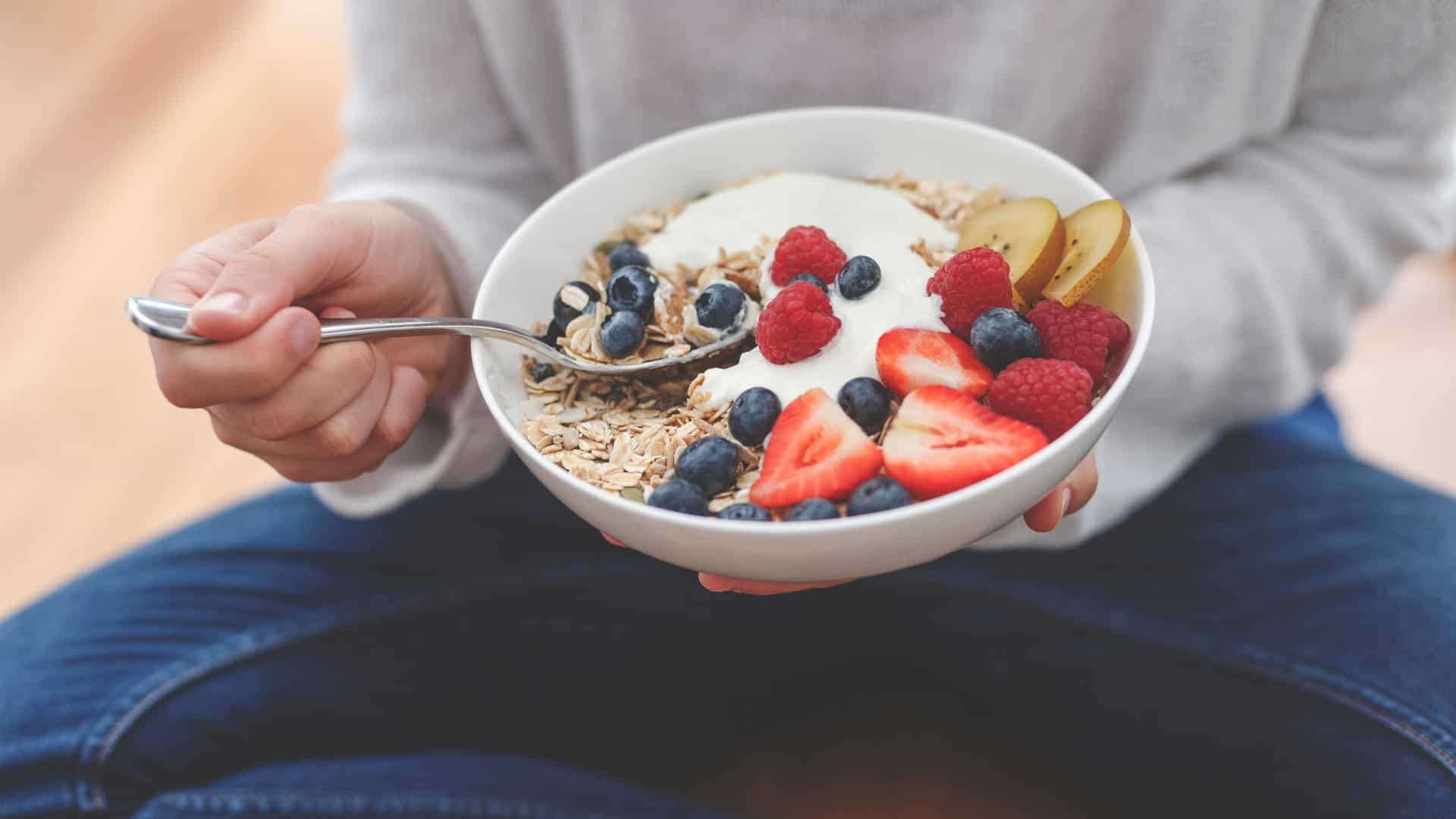 Woman sits holding a bowl of muesli topped with fresh blueberries, strawberries and kiwi fruit