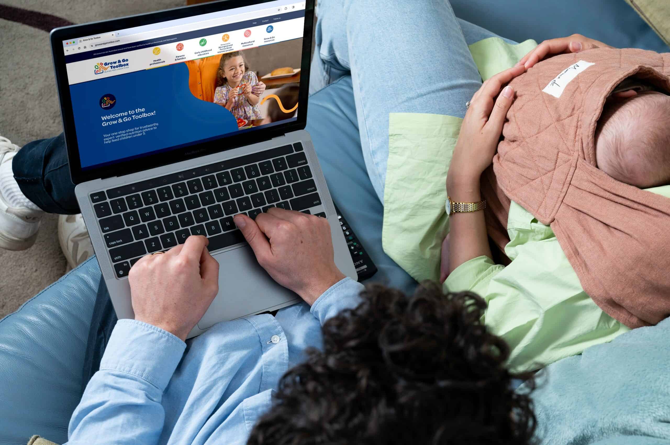 Man on laptop looking at Grow & Go Toolbox website, seating beside a woman wearing a baby carrier holding a baby