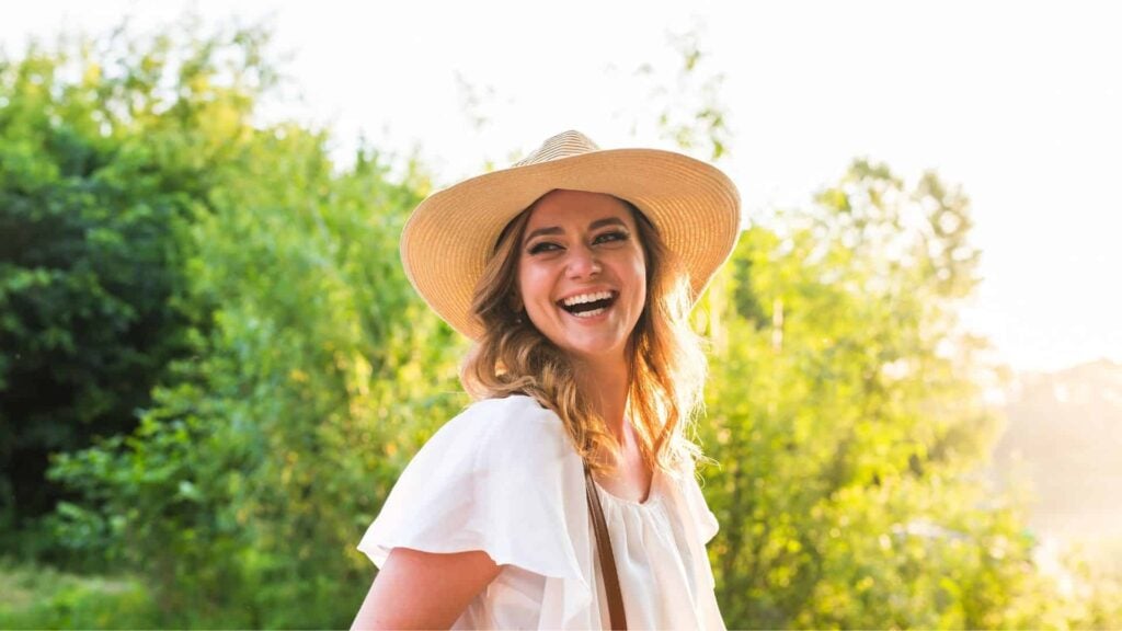 Young woman wearing straw hats stands outside in the sun laughing to someone out of sight