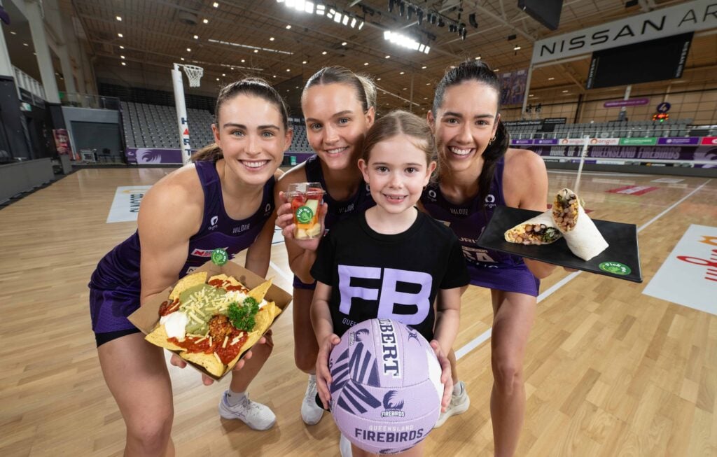 Queensland Firebirds Macy Gardner, Emily Moore and Tippah Dwan hold up healthy food options with a young fan who is holding a netball.