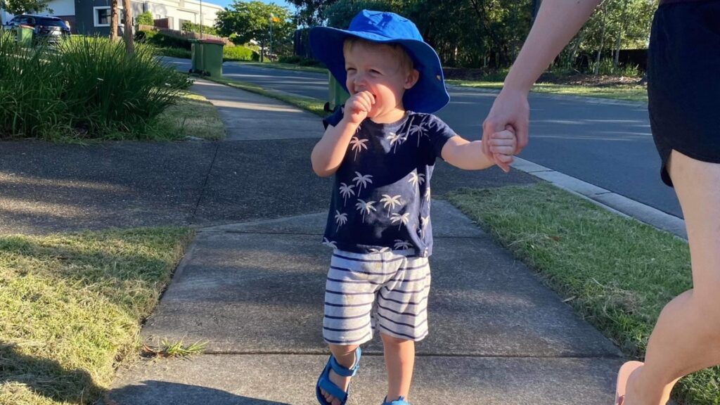 Young boy wearing blue hat walks outside holding his mother's hand