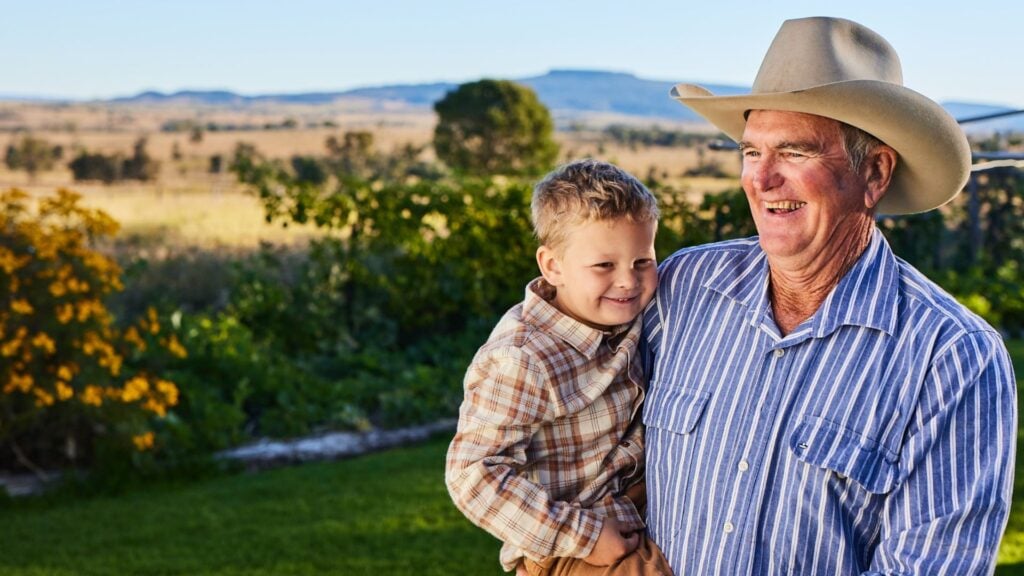 An older man is holding a young boy outside in a rural location.