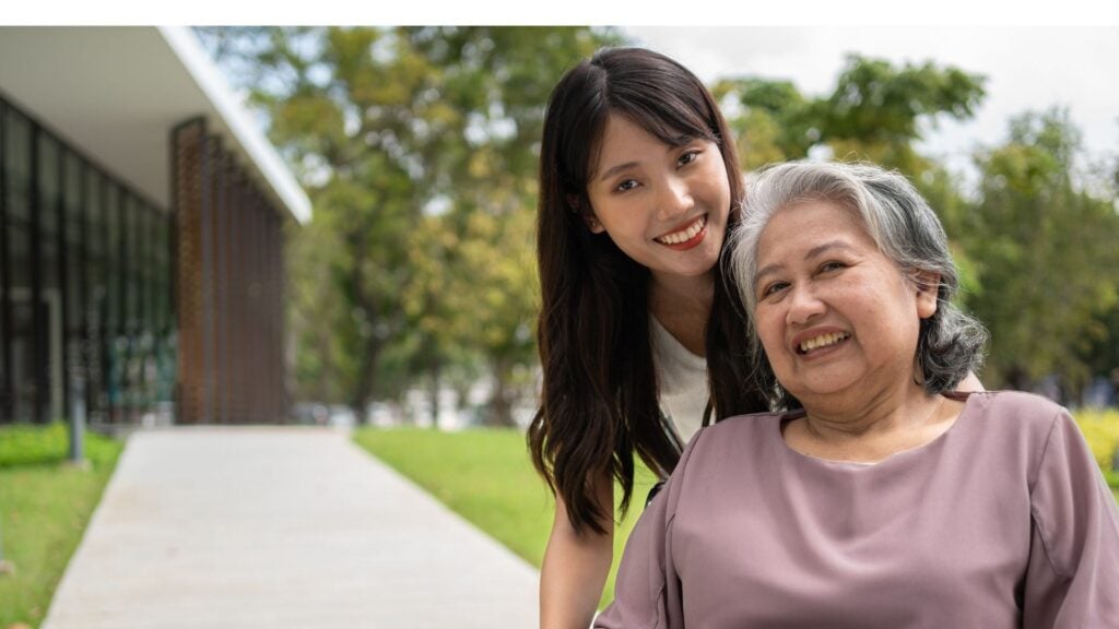 Woman in wheelchair smiles at the camera with her daughter standing behind her smiling as well