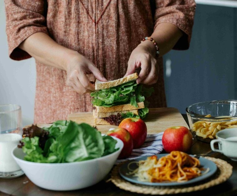 Woman preparing sandwich in kitchen with other foods also on table.