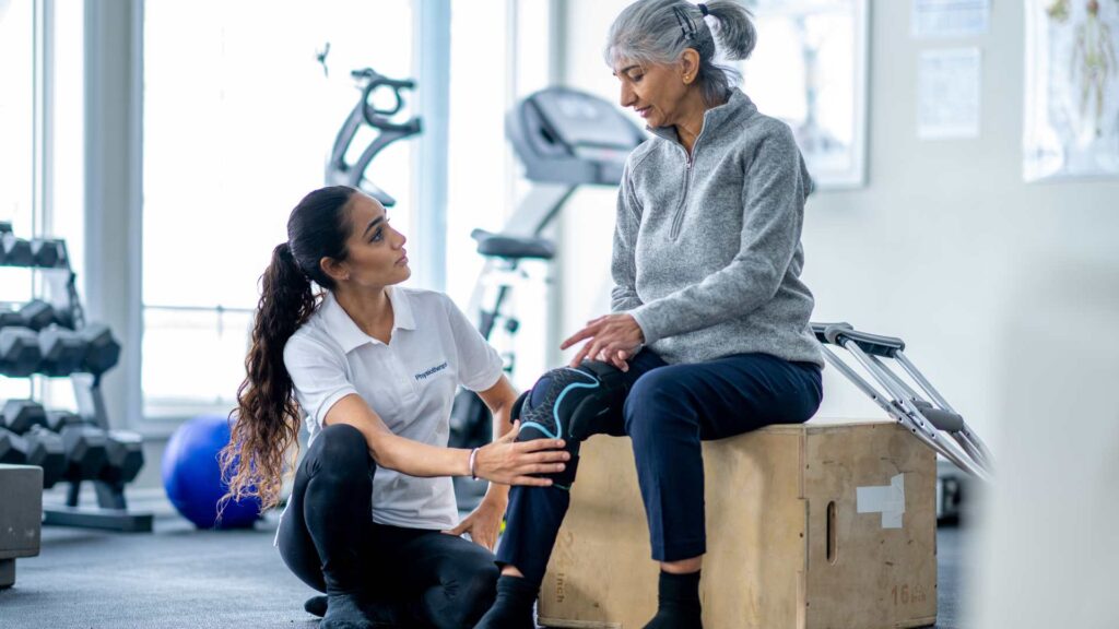 Woman wearing knee brace talks to an exercise trainer kneeling down beside her