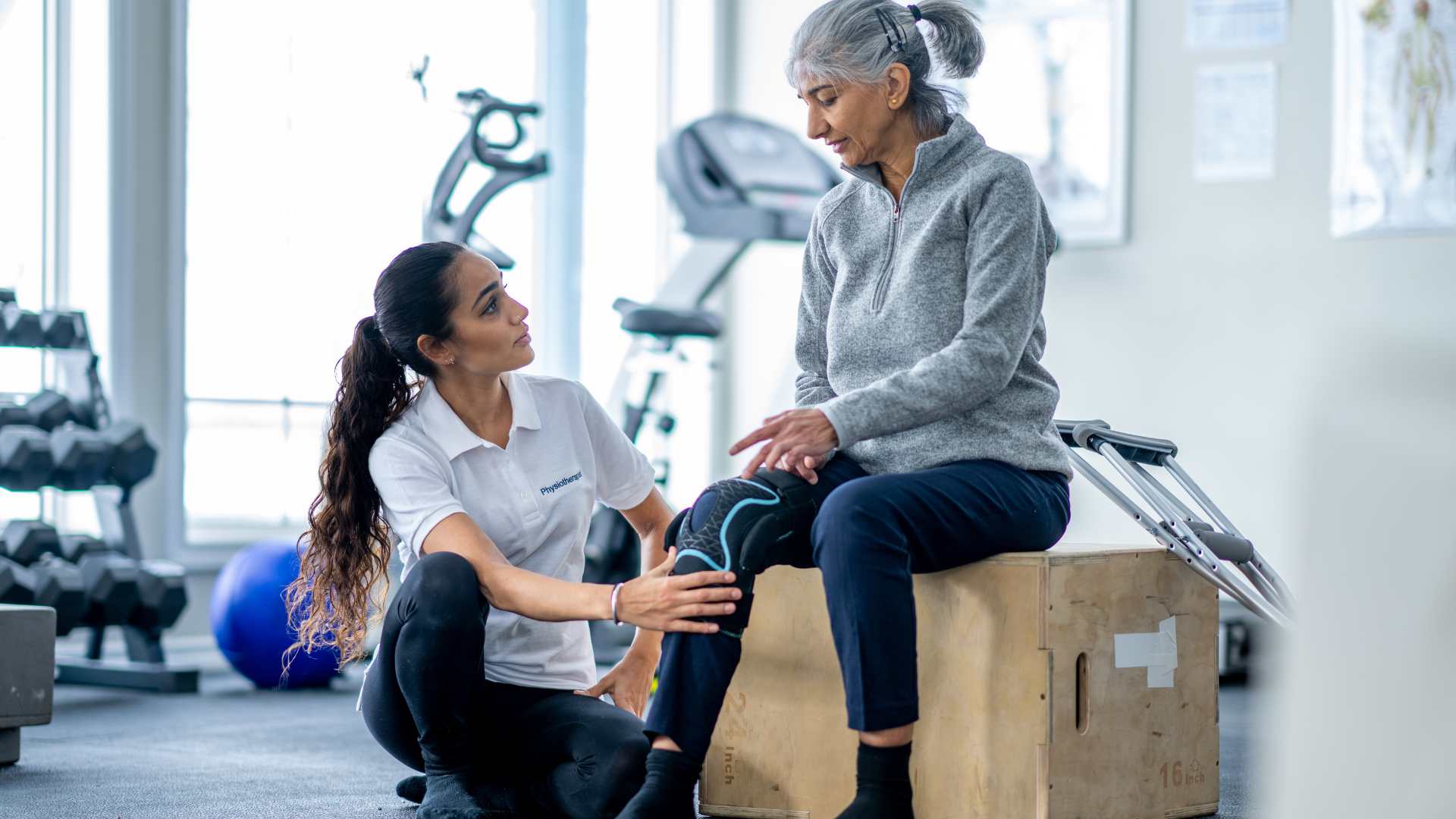 Woman wearing knee brace talks to an exercise trainer kneeling down beside her