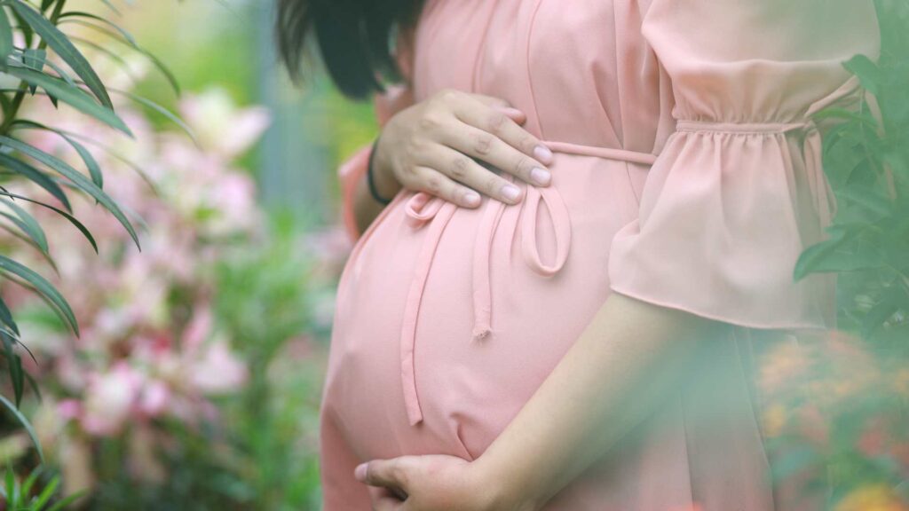 Woman in pink dress stands in the garden holding her pregnant stomach