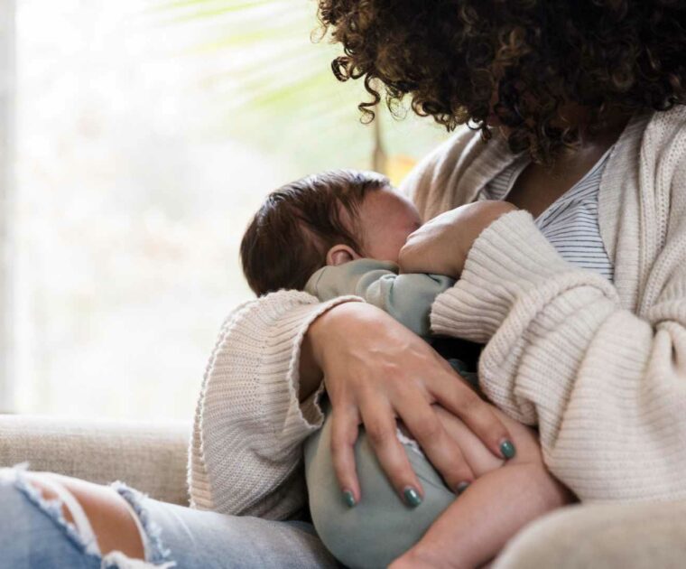 Mother with brown curly hair in striped top and cream cardigan, breastfeeding baby in green baby clothes.