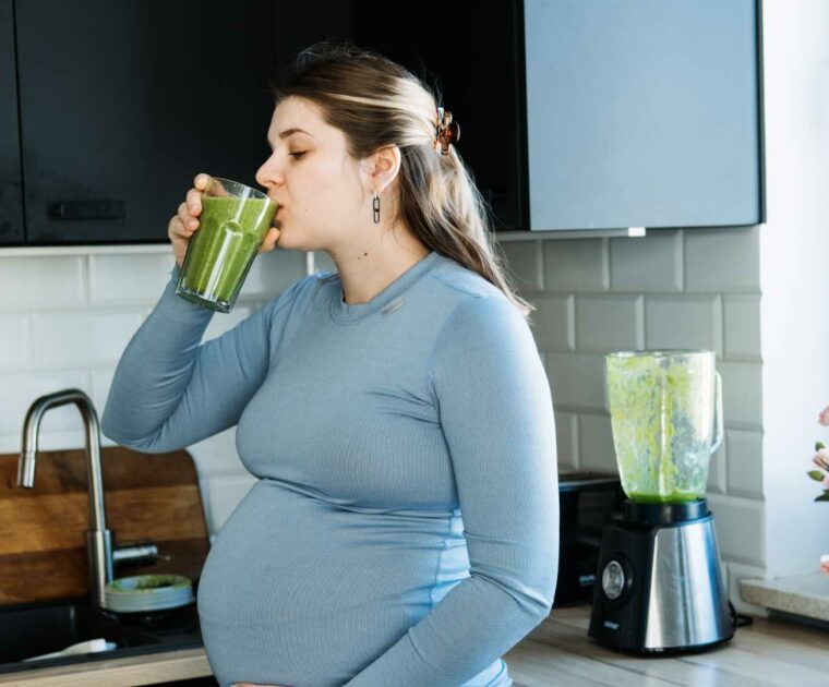 Pregnant woman in blue-grey long sleeve top in a kitchen, drinking a green smoothie out of a glass.