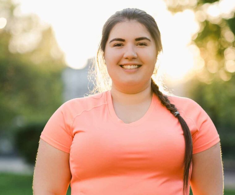 Smiling woman in orange work out t-shirt standing in an outdoor area.