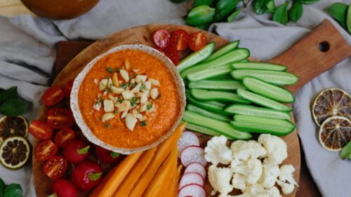 A bowl of roasted capsicum, feta and macadamia dip with freshly chopped vegetables surrounding it on a serving board