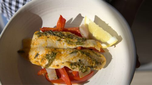 Baked fish on a plate served over cooked red capsicum, with a wedge of lemon