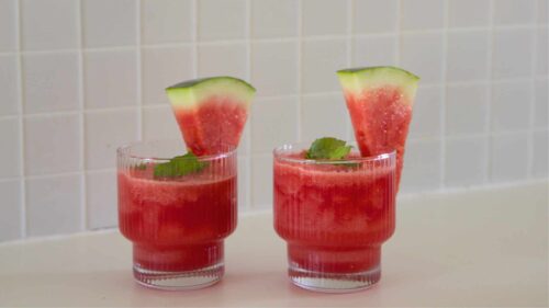 Two watermelon slushies sit on a countertop, with fresh watermelon slices to decorate them