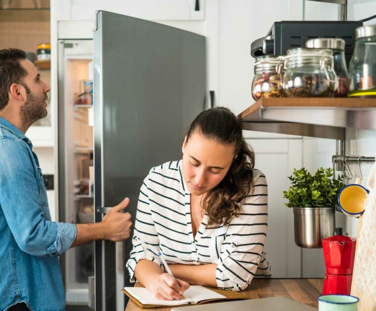 Man and woman in kitchen, with man opening fridge and woman writing in notepad on kitchen bench.