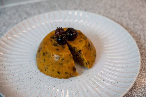 A festive Christmas pudding made with sweet potato, topped with three fresh cherries, served on a speckled white plate. A slice has been cut out, revealing the moist and rich texture of the dessert inside.