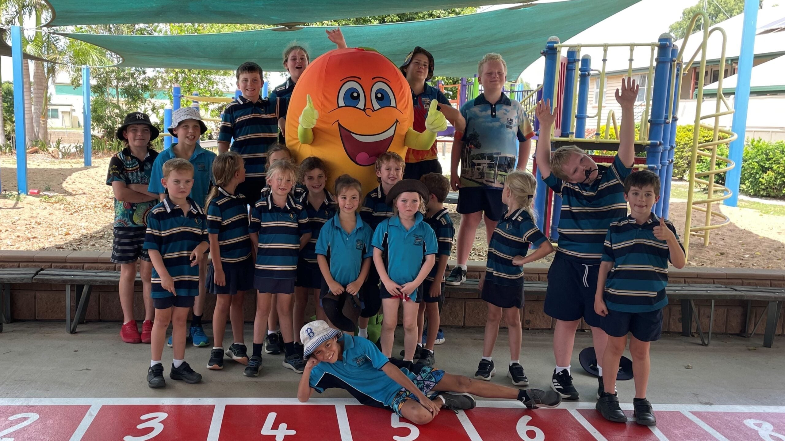 A group of school students from Clare State School stand in a playground with mascot, Kenny the Mango. 