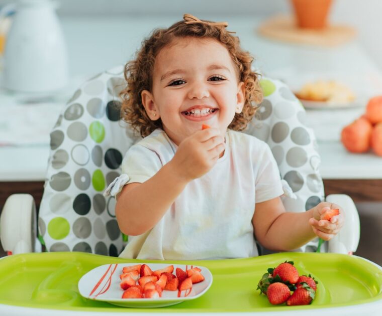 Young girl sitting on highchair holding cut piece of strawberry
