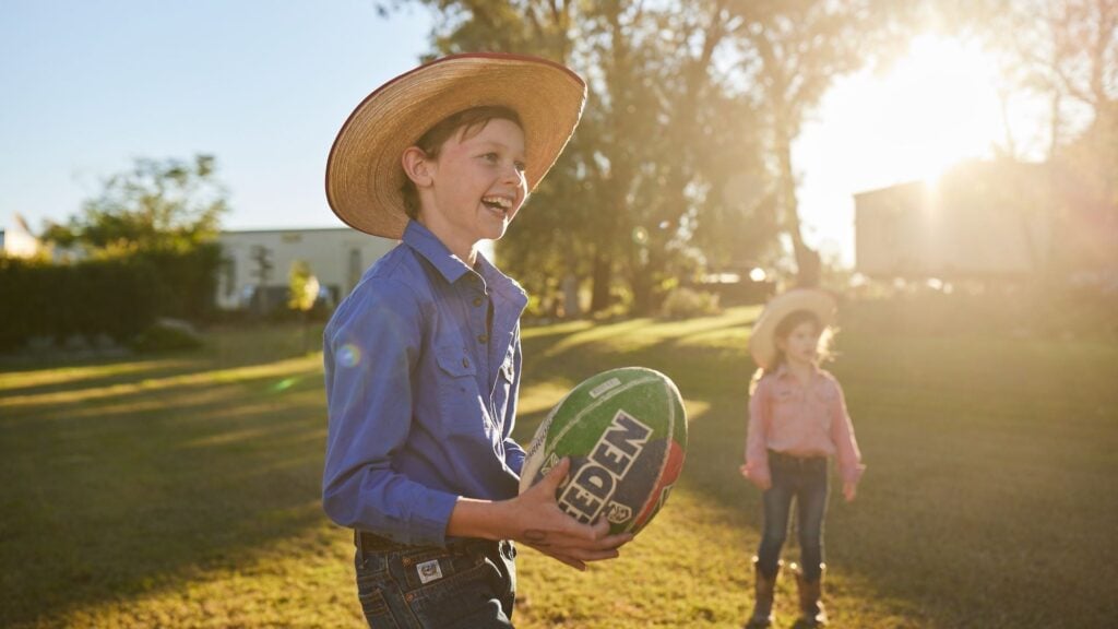 A young boy holding a football outside in a sunny yard. A young girl stands behind him.