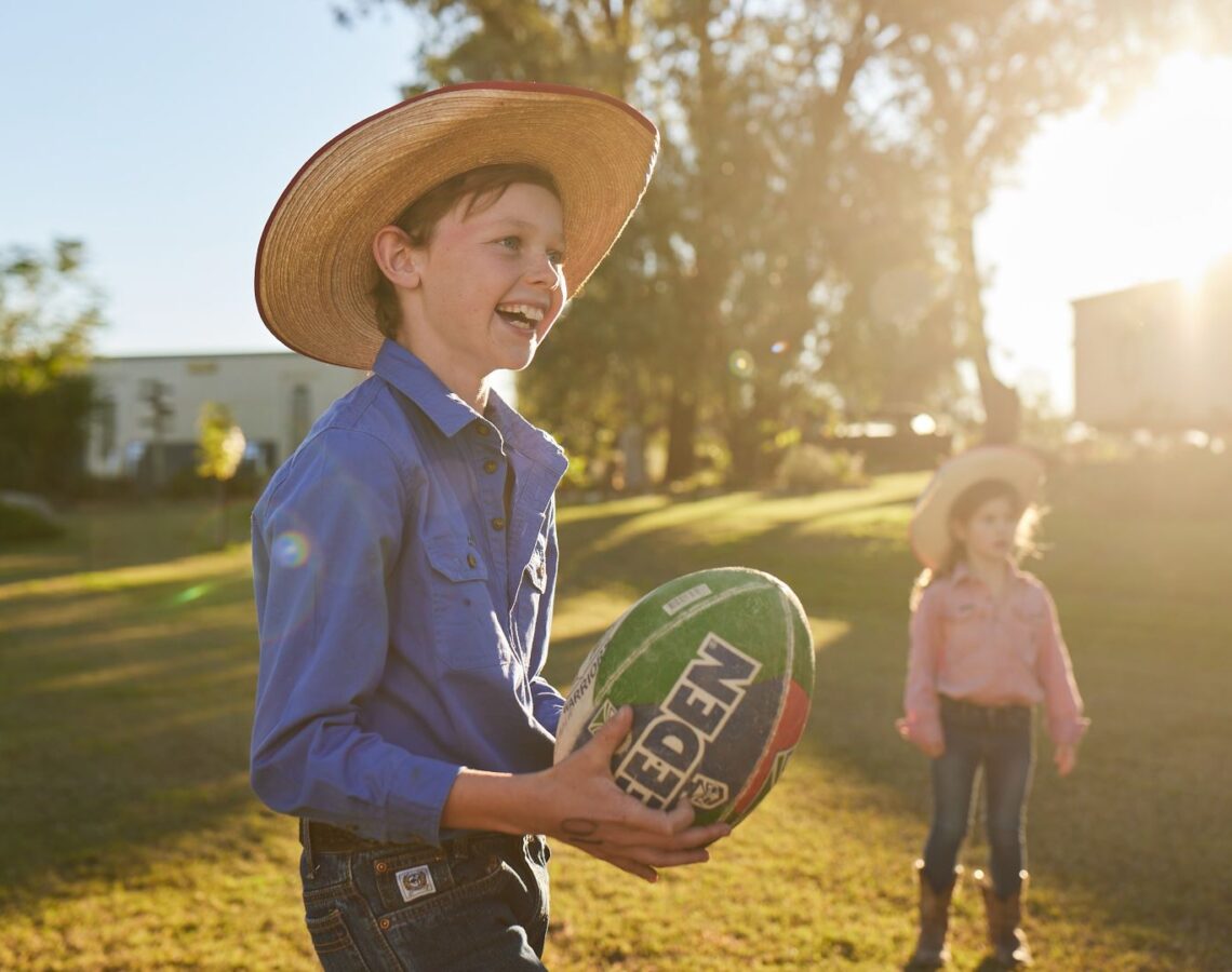 A young boy holding a football outside in a sunny yard. A young girl stands behind him.