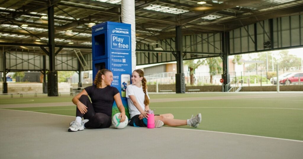 Two individuals sitting on the edge of a netball court next to a SportsBox locker. One individual is holiding a netball and the other is holding a drink bottle.
