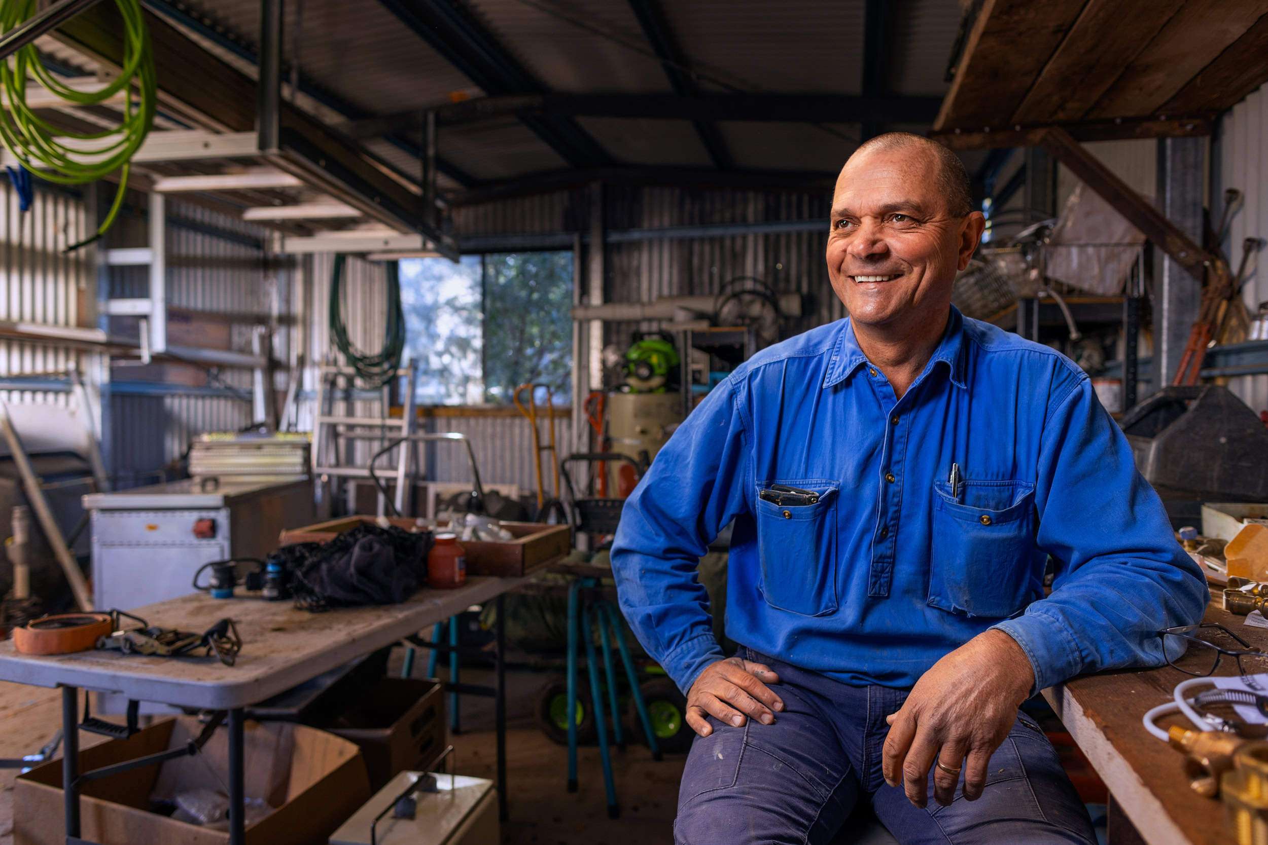 Person smiling in a workshop, sitting on a table with tools and equipment in the background. The individual is wearing a blue work shirt.