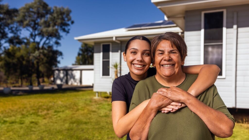 Two people smiling and embracing in front of a house with a green lawn and clear blue sky.