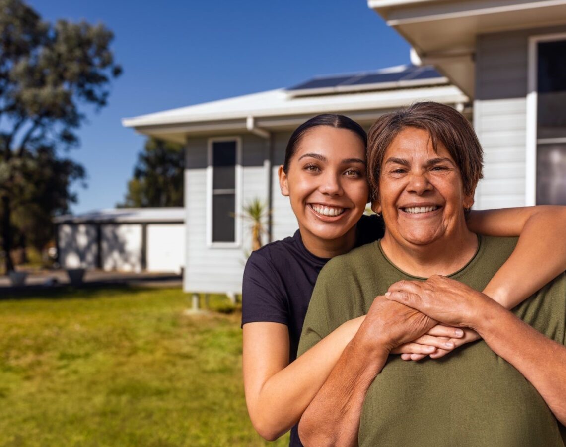 Two people smiling and embracing in front of a house with a green lawn and clear blue sky.