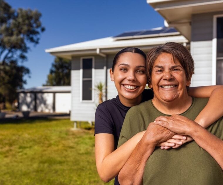 Two people smiling and embracing in front of a house with a green lawn and clear blue sky.