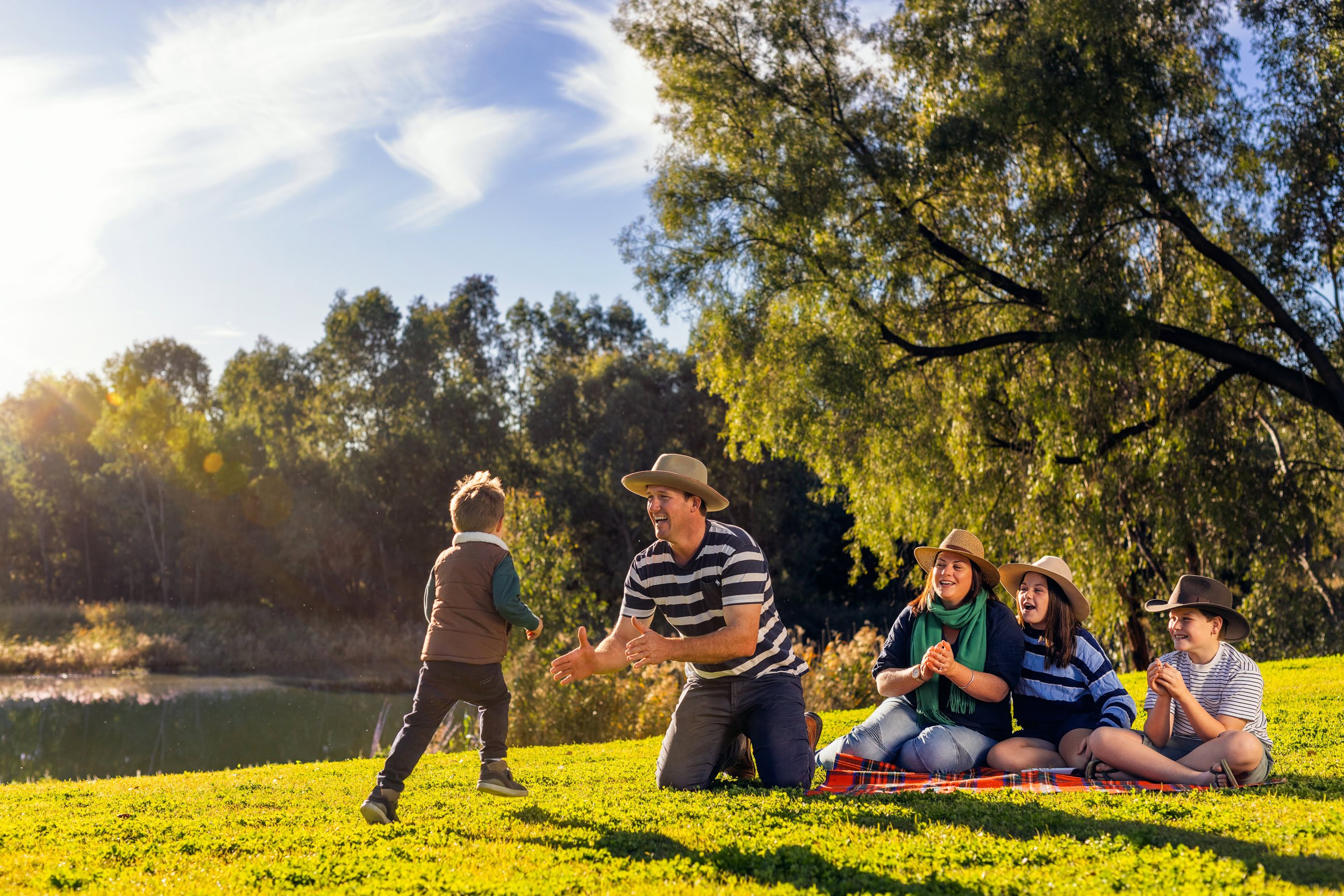 A family enjoying a sunny day outdoors, with one adult playing with a young child on the grass while three other family members sit on a blanket watching and smiling.