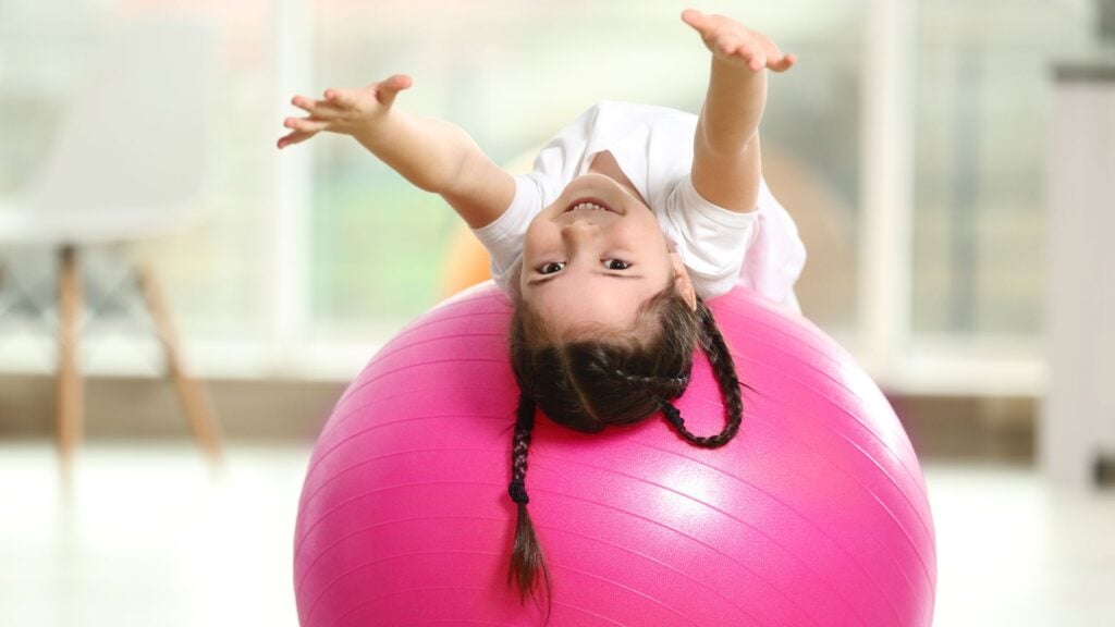 Young girl with pigtails lays on her back over pink exercise ball