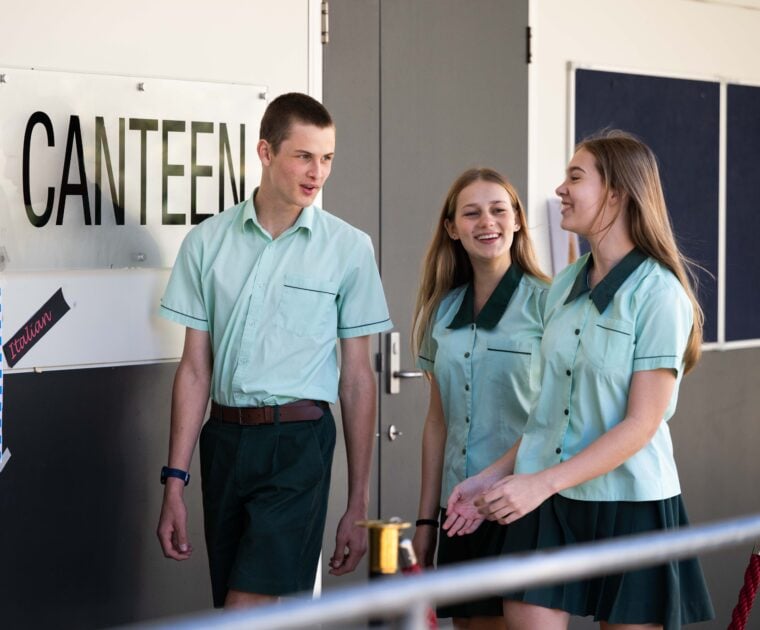 Three students in school uniform walk together down a ramp outside a canteen.