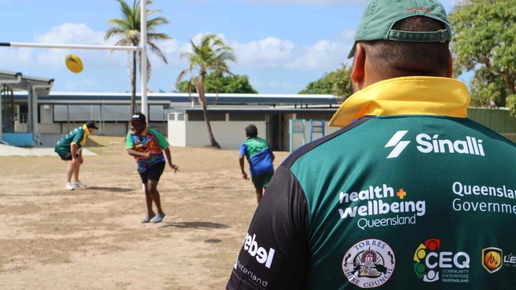 Adult male rugby players watch on as children play rugby on the field