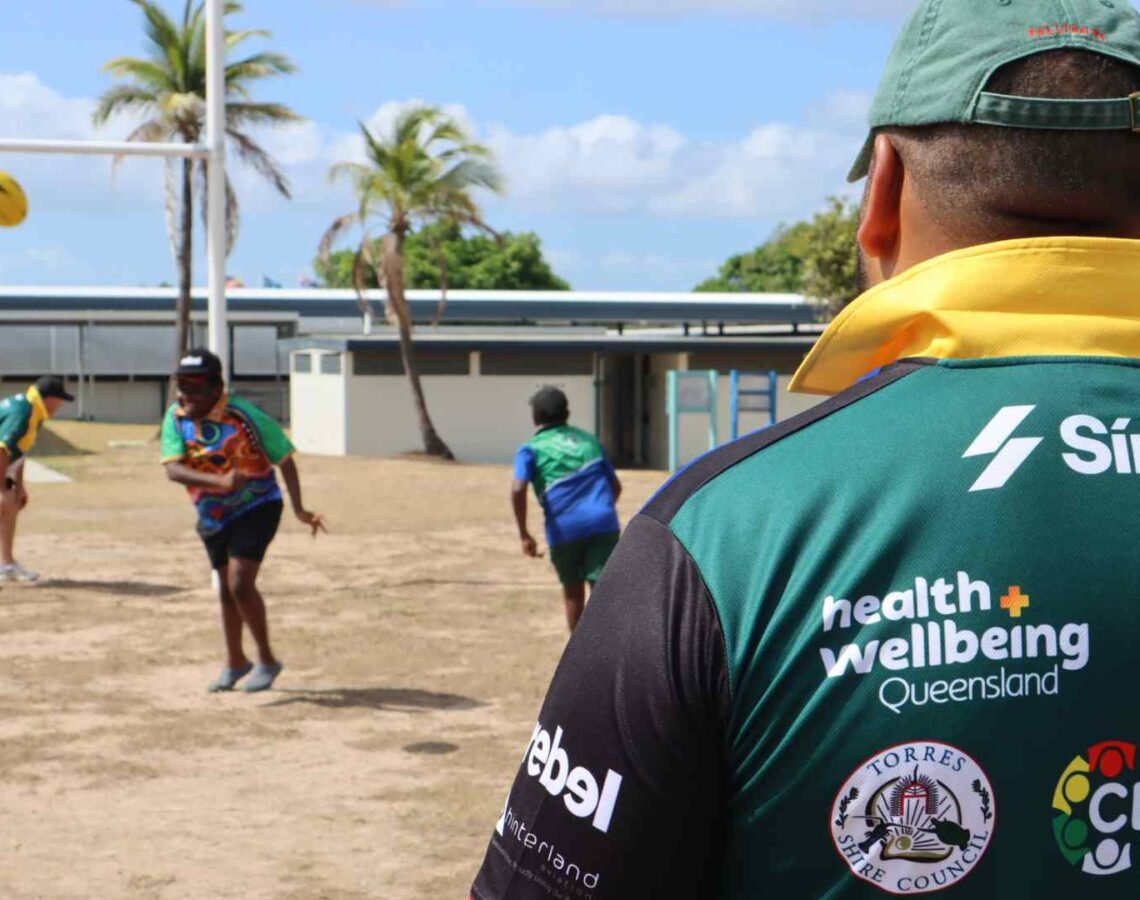 Adult male rugby players watch on as children play rugby on the field