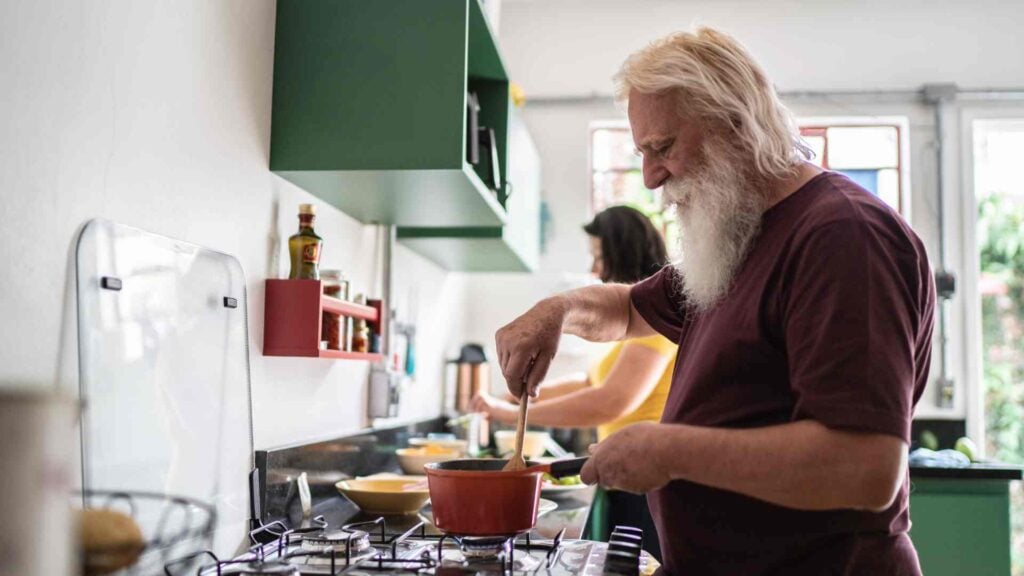 Older man stands at the stove stiring a pot while a woman in the background preps food