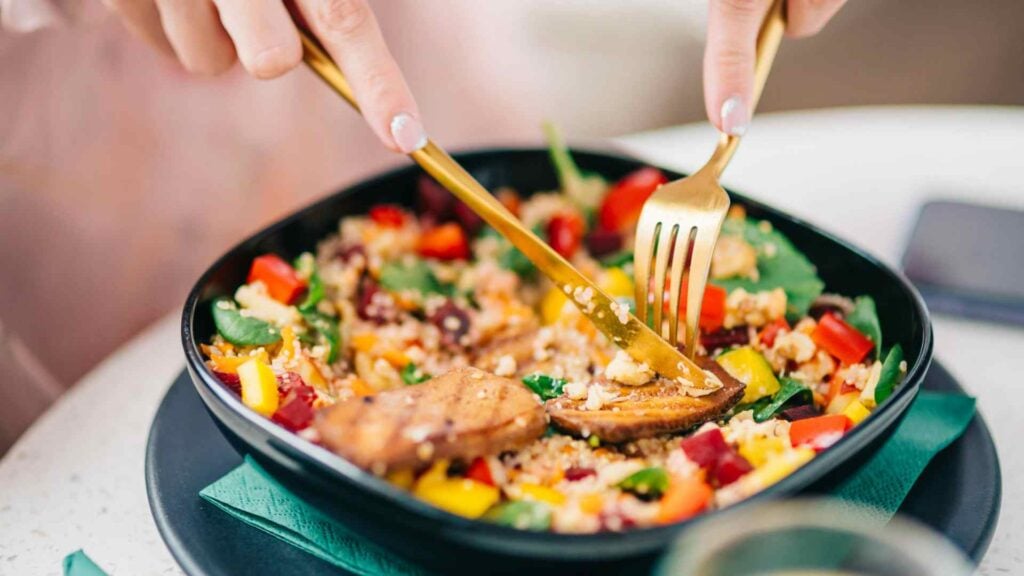 Close up on someone using a knife and fork to eat from a bowl of colourful vegetables and lean meat protein