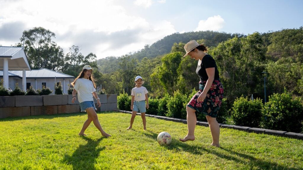 Mum kicks a soccer ball around in backyard with her 2 daughters