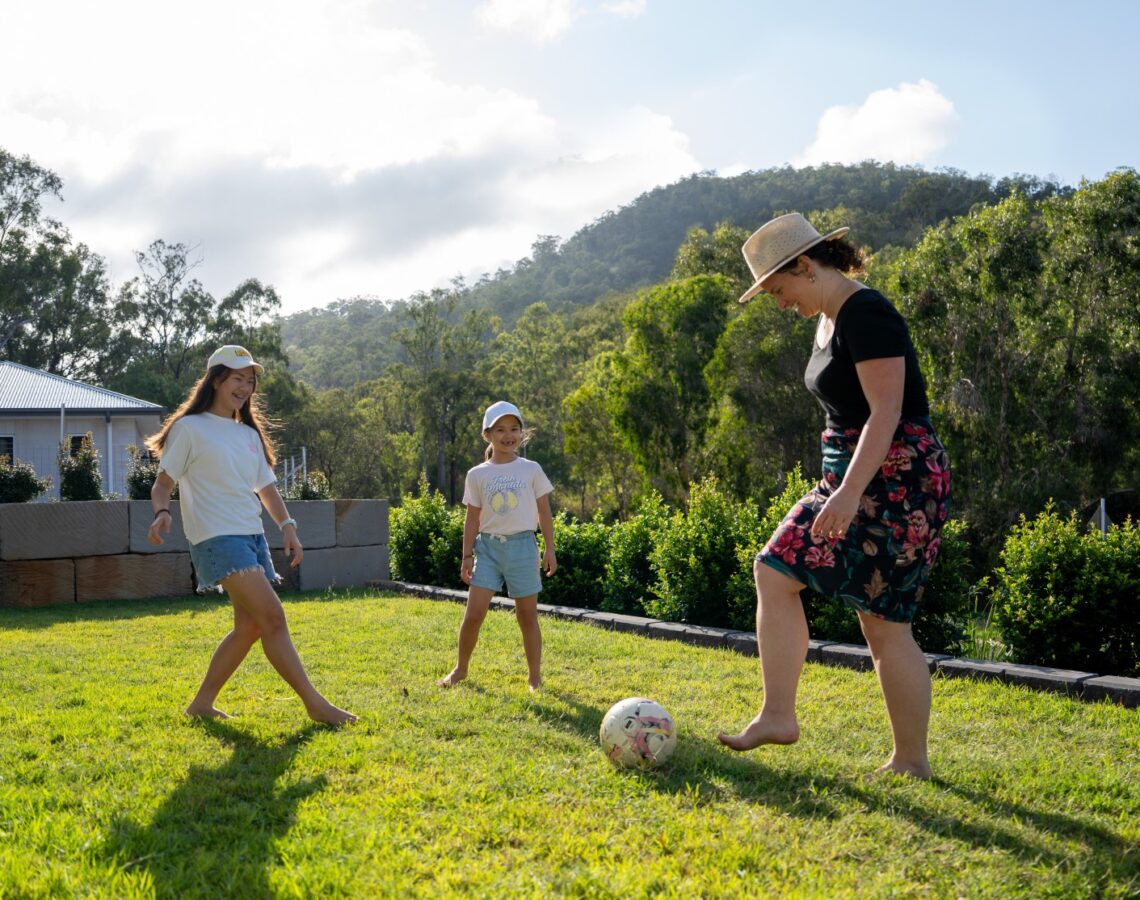 Mum kicks a soccer ball around in backyard with her 2 daughters