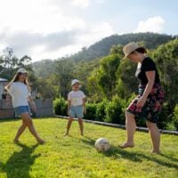 Mum kicks a soccer ball around in backyard with her 2 daughters