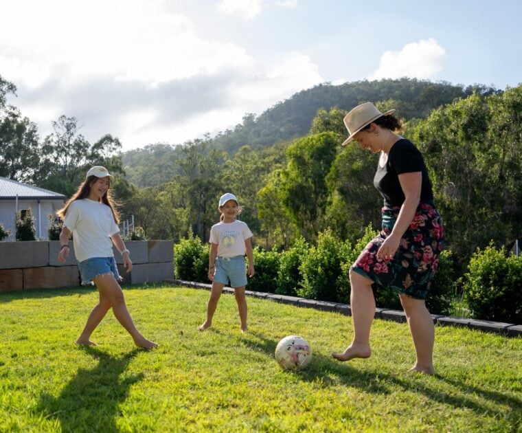 Mum kicks a soccer ball around in backyard with her 2 daughters