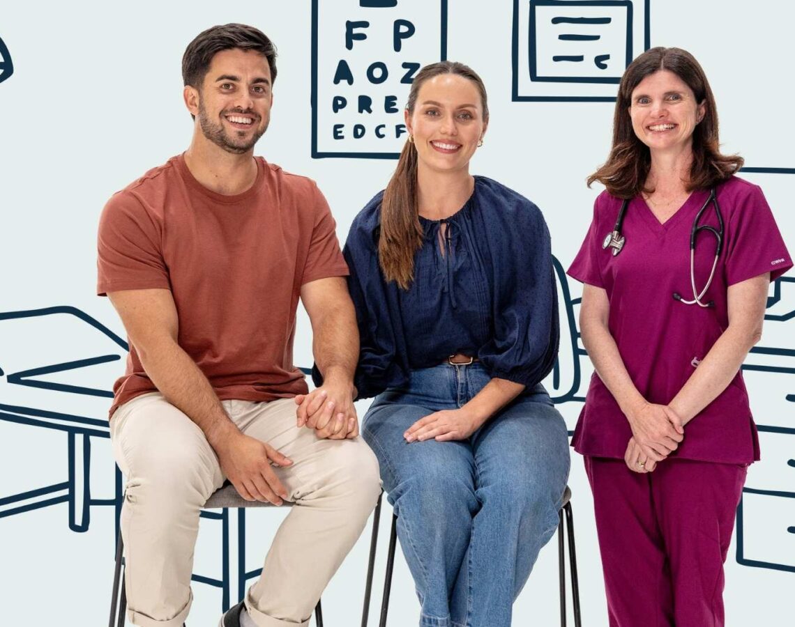 Young couple sit holding hands beside a doctor all smiling to camera